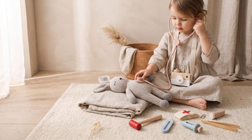 Little girl in a linen dress listening to a knitted bunny with a wooden toy stethoscope, doctor kit pieces on the rug